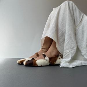 May include: A pair of brown, white, and black patterned Mary Jane style flats with a buckle closure. The shoes have a fur-like texture and are worn with a white garment. The shoes are on a gray surface.