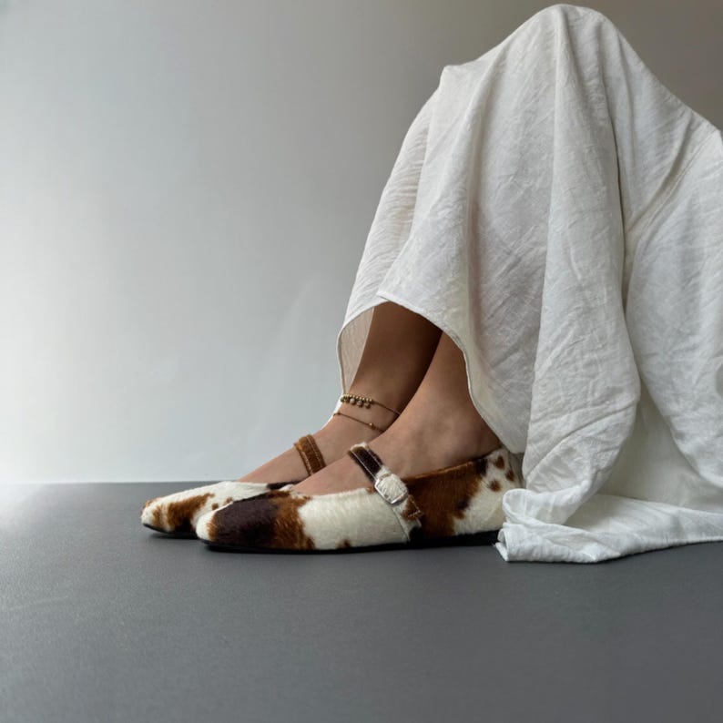 May include: A pair of brown, white, and black patterned Mary Jane style flats with a buckle closure. The shoes have a fur-like texture and are worn with a white garment. The shoes are on a gray surface.