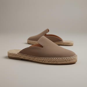 May include: Taupe-colored espadrille mules with a slip-on design. The shoes feature a woven jute sole and a smooth upper. The second mule is slightly out of focus in the background. The shoes are a neutral color, suitable for various outfits.