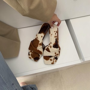 May include: A pair of open-toe sandals with a cowhide pattern. The sandals feature a brown and white spotted design, with a square toe and a strap across the top of the foot. The sandals are held by a person.