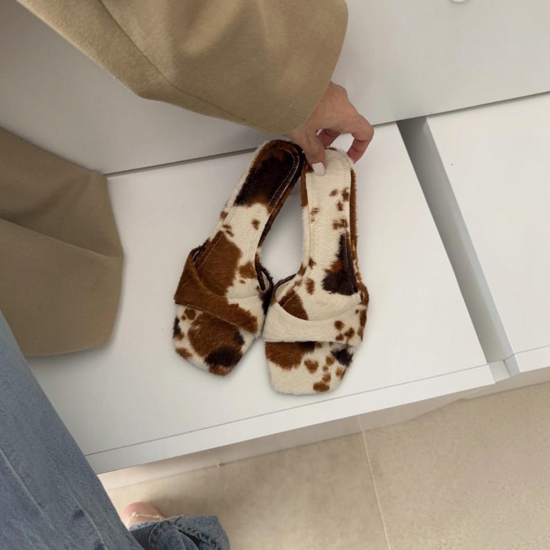 May include: A pair of open-toe sandals with a cowhide pattern. The sandals feature a brown and white spotted design, with a square toe and a strap across the top of the foot. The sandals are held by a person.