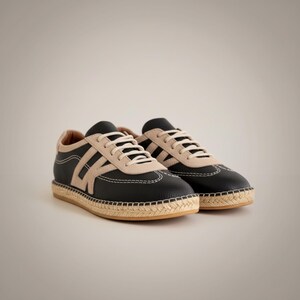May include: A pair of black trainers with beige detailing and white laces. The shoes feature a woven sole and stitched design. These trainers are a casual style, suitable for everyday wear.