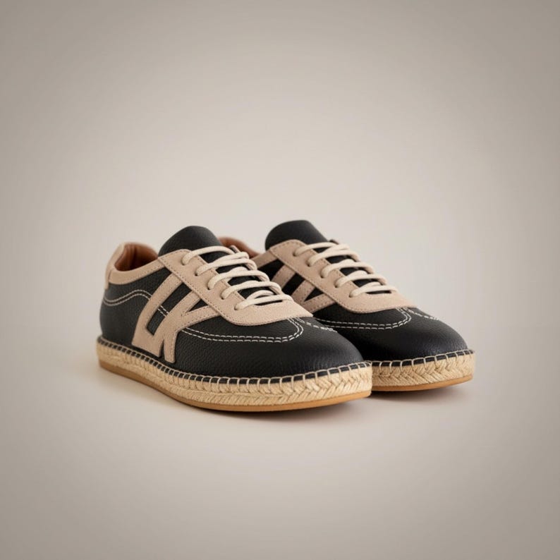 May include: A pair of black trainers with beige detailing and white laces. The shoes feature a woven sole and stitched design. These trainers are a casual style, suitable for everyday wear.