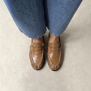 May include: A pair of tan leather loafers featuring a classic penny slot strap across the vamp, visible stitching along the moccasin-style toe, and a smooth finish, worn with blue denim jeans that extend down to the top of the footwear.