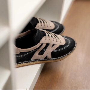 May include: A pair of black and beige trainers with white stitching and beige laces. The trainers feature a suede accent and a textured sole. The shoes are on a white shelf.