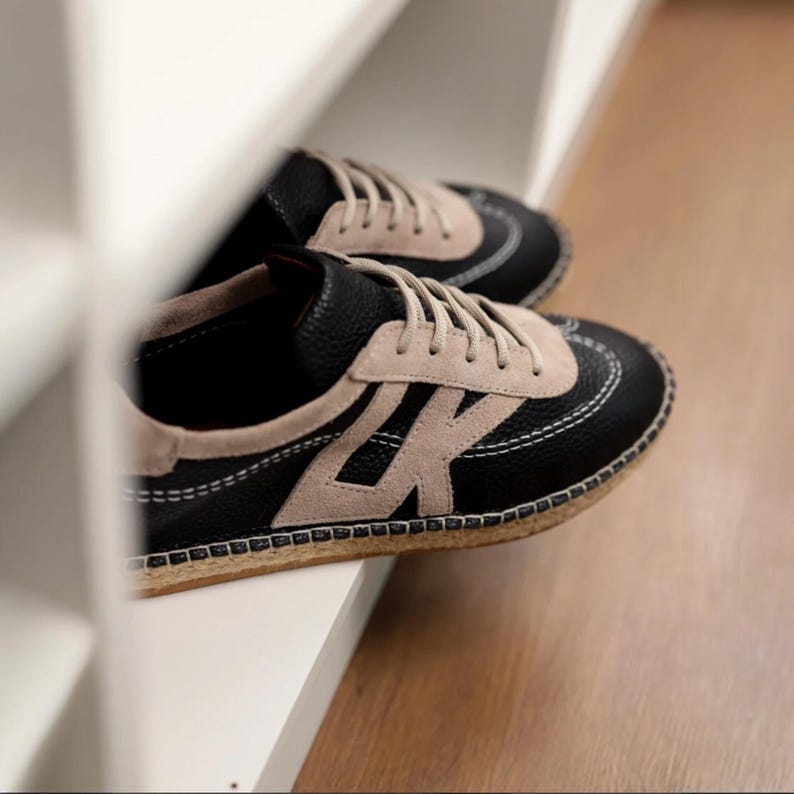 May include: A pair of black and beige trainers with white stitching and beige laces. The trainers feature a suede accent and a textured sole. The shoes are on a white shelf.