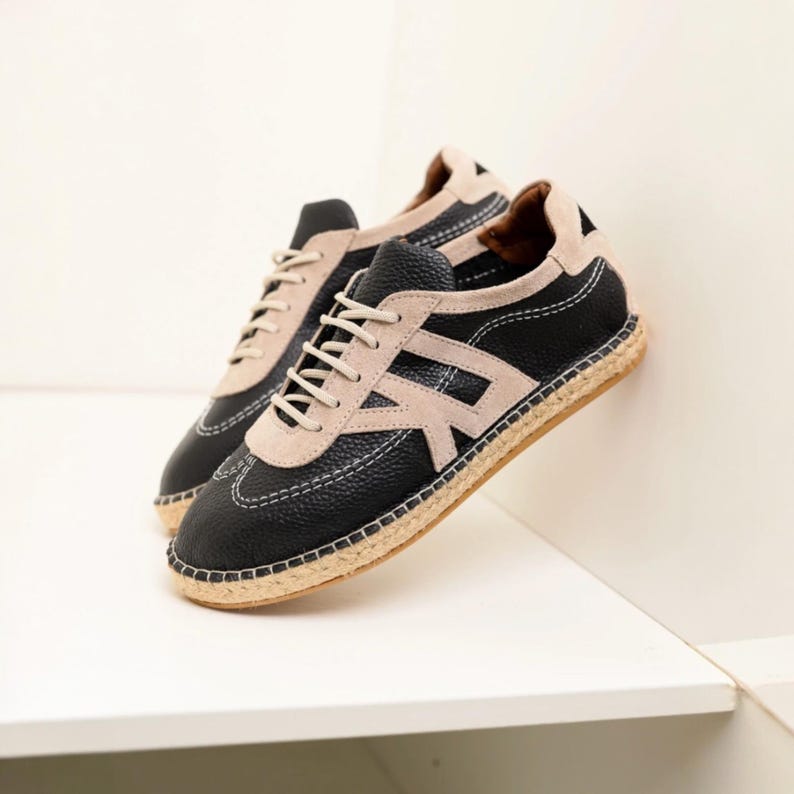 May include: Black and beige sneakers with a textured leather upper and a beige suede accent. The sneakers feature a beige "R" design on the side, white stitching, and a woven sole. The laces are beige.