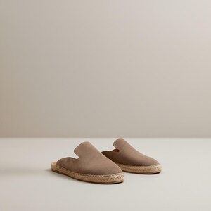 May include: A pair of taupe-colored slip-on espadrilles. The shoes have a suede-like upper and a woven jute sole. The design features a rounded toe and a low-profile heel, suitable for casual wear. The neutral color makes them versatile for various outfits.