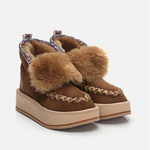Könnte beinhalten: Braune Wildleder-Plateau-Stiefel mit beigefarbener Sohle und hellbraunem Fellbesatz. Die Stiefel haben dekorative Nähte und gemusterte Stoffdetails. Modische Schuhe.