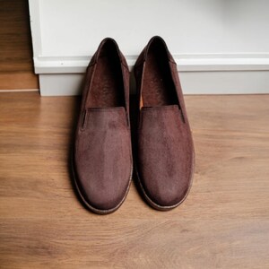 Puede incluir: Un par de zapatos sin cordones de ante marr&oacute;n. Los zapatos tienen una puntera redondeada y un perfil bajo, con un dise&ntilde;o sencillo. Los zapatos est&aacute;n hechos de un material suave y texturizado y son adecuados para ropa informal.