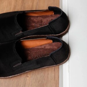 Puede incluir: Un par de zapatos negros sin cordones con plantillas marrones. Los zapatos tienen una punta redondeada y un perfil bajo. Las plantillas son de color marr&oacute;n contrastante. Los zapatos est&aacute;n colocados sobre una superficie de madera.