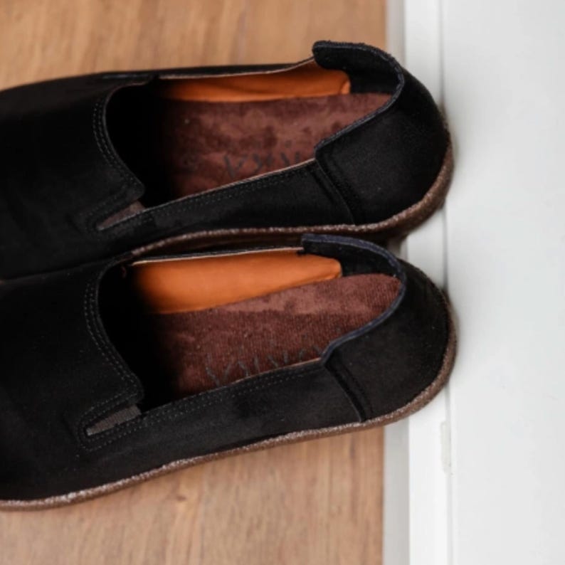 Puede incluir: Un par de zapatos negros sin cordones con plantillas marrones. Los zapatos tienen una punta redondeada y un perfil bajo. Las plantillas son de color marr&oacute;n contrastante. Los zapatos est&aacute;n colocados sobre una superficie de madera.