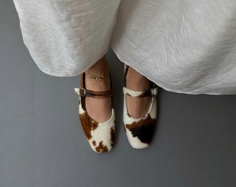 May include: A pair of cowhide print Mary Jane flats. The shoes feature a brown leather strap and buckle closure. The uppers are a mix of brown and white, creating a unique pattern. The shoes are set against a gray background.