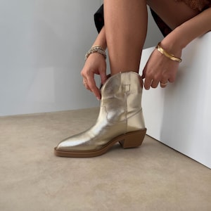 May include: A metallic gold cowboy boot with a low heel and a rounded toe. The boot has a smooth, shiny finish and a brown sole. The person is wearing bracelets.