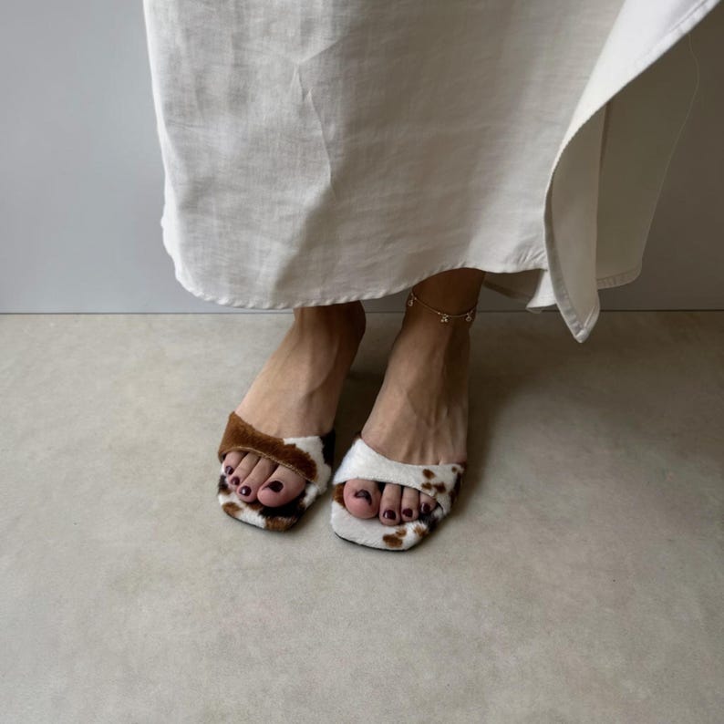 May include: A pair of square-toed slip-on sandals with a cowhide pattern. One sandal features brown fur, while the other has white fur with brown spots. The sandals are worn with a long, flowing white dress.