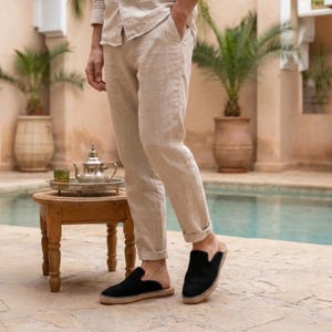 May include: Black suede slip-on shoes with a woven jute sole. The shoes are worn with beige linen pants and a matching button-down shirt. The setting is outdoors, near a pool, with a small wooden table and a silver tea set.