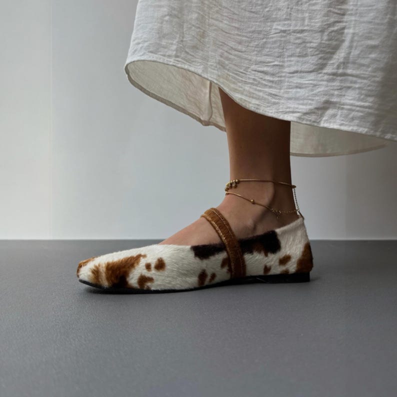 May include: Flat ballet-style shoe with a cowhide pattern in brown and white. The shoe has a brown strap across the top and a black sole. A gold ankle bracelet is also visible.