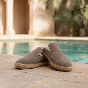 May include: A pair of olive green suede slip-on espadrilles. The shoes have a rounded toe and a woven jute sole. The background features a pool and a building with columns.