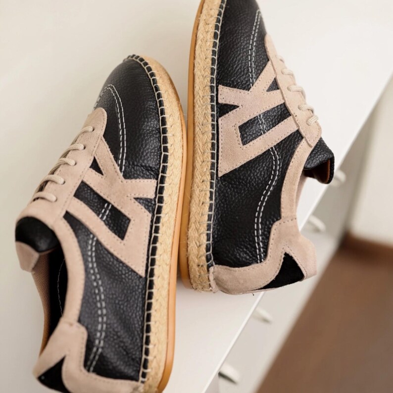 May include: A pair of black and beige sneakers with a textured leather upper and a woven jute sole. The sneakers feature a beige "V" design on the sides and white stitching details. The soles are approximately 2.5 cm thick.
