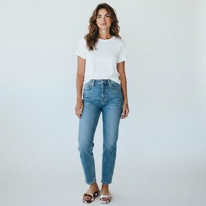 May include: A woman wearing a white crew neck t-shirt and light blue denim jeans. The jeans have a high waist and a straight leg fit. She is also wearing patterned sandals. The background is plain white.