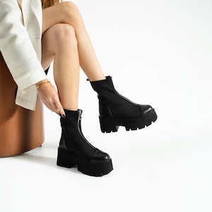 Puede incluir: Botines negros con plataforma y cierre de cremallera. Las botas tienen una suela gruesa y una puntera redondeada. La parte superior de la bota es de un material liso y mate. Las botas las lleva una persona sentada.