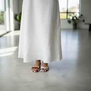 May include: A long, flowing white dress with a simple design. The hem of the dress falls to the ankles. The person is wearing sandals with a brown and white pattern.