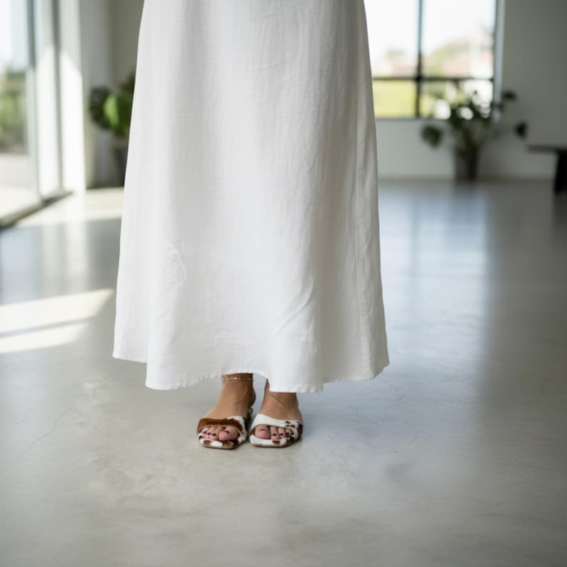 May include: A long, flowing white dress with a simple design. The hem of the dress falls to the ankles. The person is wearing sandals with a brown and white pattern.