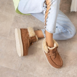 Könnte beinhalten: Braune Wildleder-Ankle Boots mit cremefarbenem Kunstfellbesatz und einer dicken, cremefarbenen Sohle. Die Stiefel haben ein Schnürdesign und werden mit hellblauen Jeans getragen.