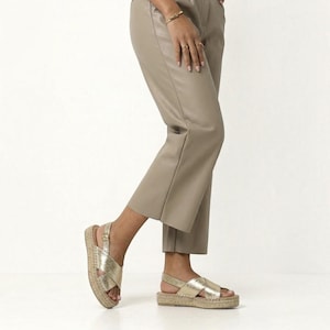 May include: A pair of metallic gold espadrille sandals featuring crisscross straps, an adjustable ankle strap with a buckle, and a woven jute platform sole, worn with cropped taupe trousers that have a smooth, matte finish.