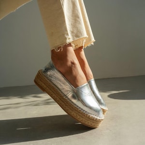 Può includere: Scarpe espadrillas metallizzate argentate con suola in juta intrecciata. Le scarpe hanno un design slip-on e una punta arrotondata. L'immagine mostra le scarpe indossate con pantaloni color crema.