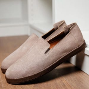 Puede incluir: Un par de zapatos sin cordones de ante marr&oacute;n claro con suela marr&oacute;n oscuro. Los zapatos se exhiben sobre una superficie de madera, con una estanter&iacute;a blanca al fondo. Dise&ntilde;o cl&aacute;sico y minimalista.