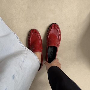 May include: A pair of red leather loafers featuring a gathered, pleated toe design and a classic penny slot strap across the vamp. The interior footbed is black and displays the brand name MUPA in white lettering. The shoes have a low profile.