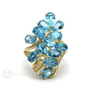 Può includere: Un anello dorato con un gruppo di pietre preziose ovali blu chiaro. L'anello ha un design floreale unico con pietre di accento più piccole. L'anello è posto su uno sfondo bianco.