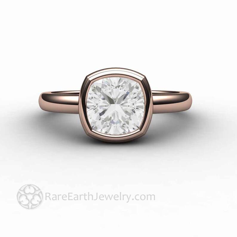 May include: A rose gold engagement ring featuring a cushion-cut diamond set in a bezel setting. The ring has a plain band and a smooth finish.
