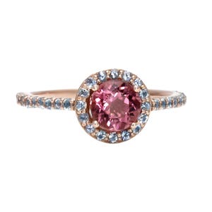 May include: A rose gold ring with a pink oval gemstone surrounded by a halo of smaller blue gemstones. The band is also set with smaller blue gemstones.