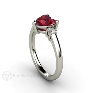 May include: A heart-shaped ruby gemstone ring with two smaller diamond accents set in a white gold band.