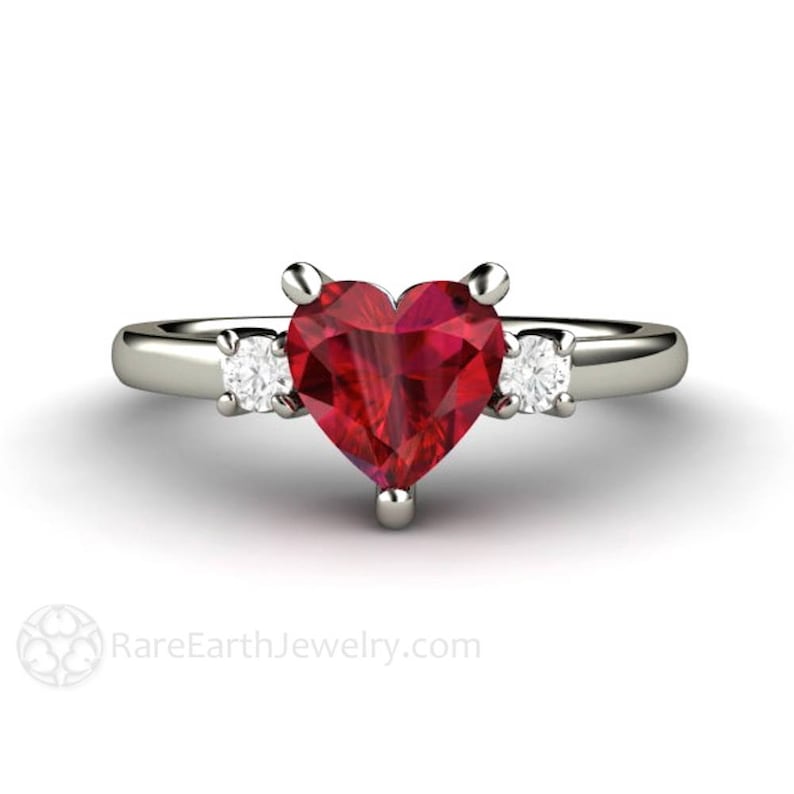 May include: A heart-shaped ruby engagement ring with two smaller diamond accents on either side of the center stone. The ring is set in white gold.