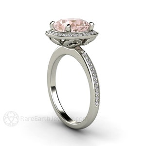 May include: A delicate engagement ring with a pale pink oval-shaped center stone surrounded by a halo of smaller diamonds. The band is set with a row of smaller diamonds.