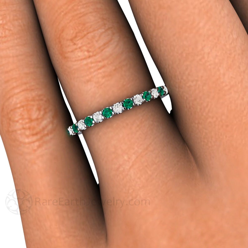 K&ouml;nnte beinhalten: Ein filigraner Ring mit abwechselnd Smaragd- und Diamant-Edelsteinen. Der Ring ist in einem silbernen Band gefasst und wird an einem Finger gezeigt. Die Edelsteine sind klein und rund und bilden ein durchgehendes Muster.