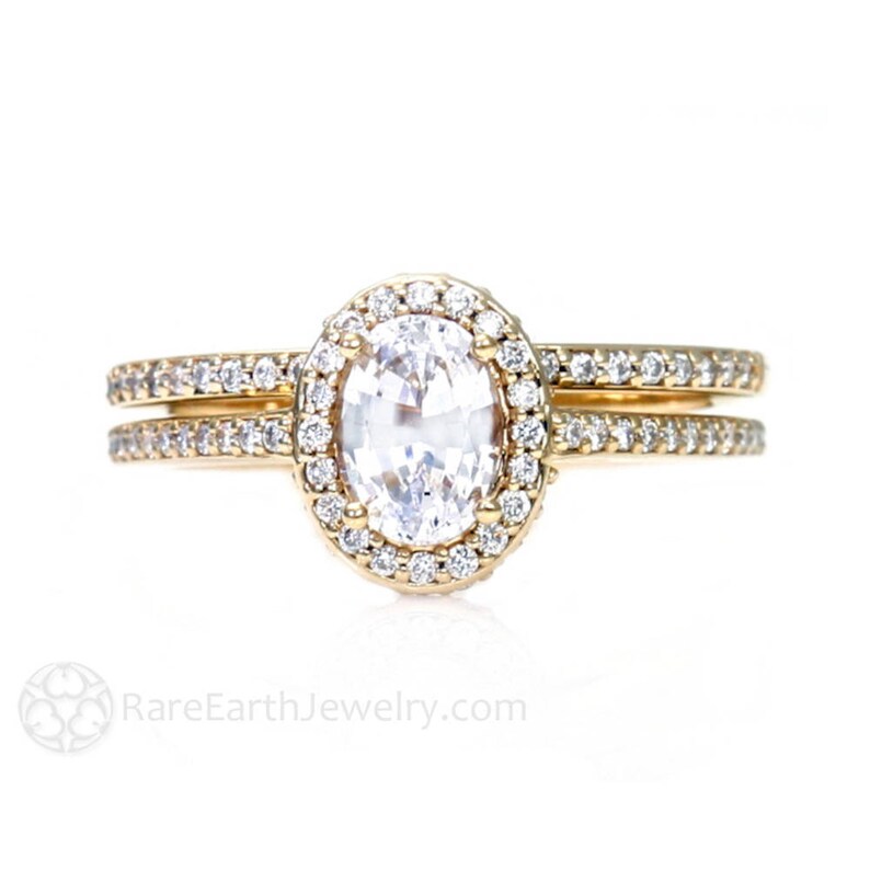 May include: A gold engagement ring set with a large oval-shaped center stone surrounded by a halo of smaller diamonds. The band is also set with smaller diamonds.