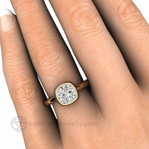 May include: A gold engagement ring with a square cushion-cut diamond set in a bezel setting. The ring is on a finger.