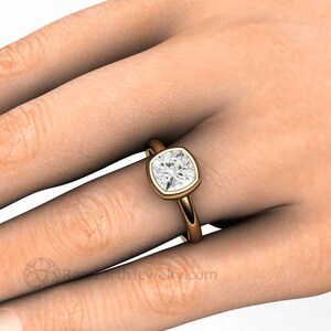 May include: A gold engagement ring featuring a cushion-cut diamond set in a bezel setting. The ring is worn on a model's finger.