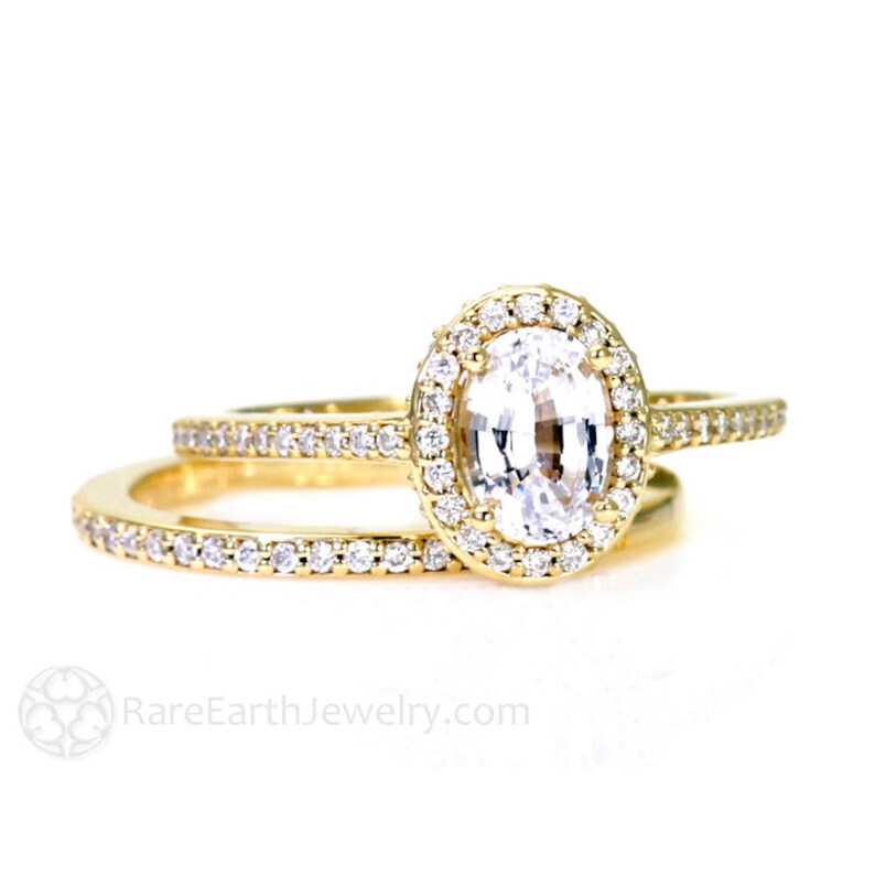 May include: A gold engagement ring set with a large oval-shaped white sapphire surrounded by a halo of smaller diamonds. The matching wedding band is also set with diamonds.