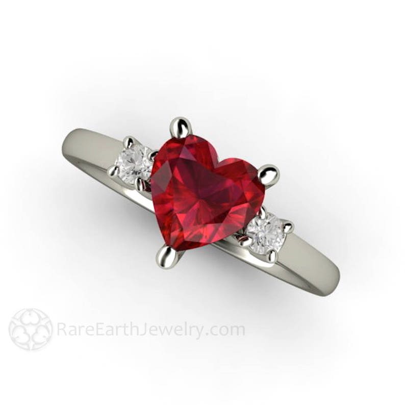 May include: A heart-shaped ruby engagement ring set in white gold with two smaller diamond accents.