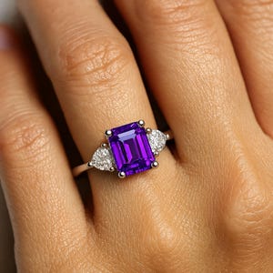 Può includere: Un anello d'argento con una grande pietra preziosa rettangolare viola scuro al centro, affiancata da due pietre preziose chiare più piccole a forma di pera. L'anello è posto su uno sfondo color carne.