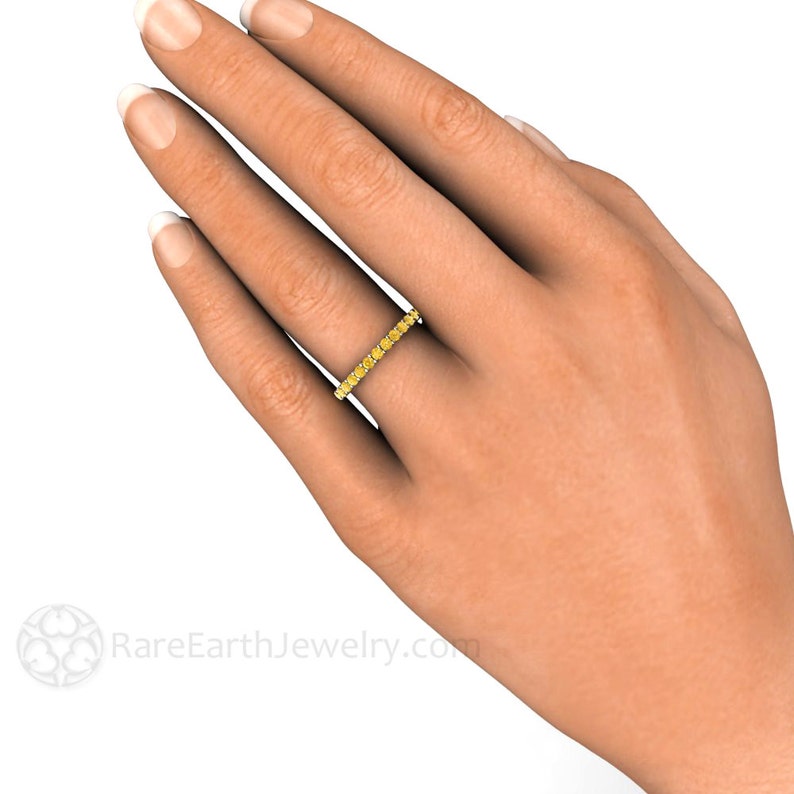 May include: A yellow gold band ring with a row of small, round, yellow gemstones. The ring is on a person's finger.