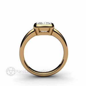 May include: A gold solitaire engagement ring with a bezel setting. The ring features a square-shaped diamond.