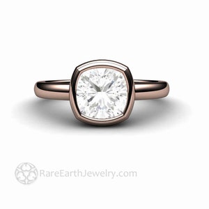 May include: A rose gold engagement ring featuring a cushion-cut diamond set in a bezel setting. The ring has a plain band and a square-shaped diamond.