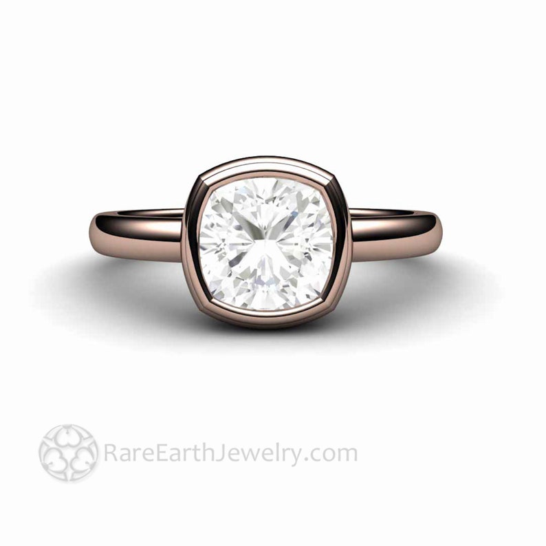May include: A rose gold engagement ring featuring a cushion-cut diamond set in a bezel setting. The ring has a plain band and a square-shaped diamond.