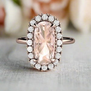 May include: A rose gold engagement ring with a large, rectangular, light pink gemstone surrounded by a halo of smaller, round, white gemstones.
